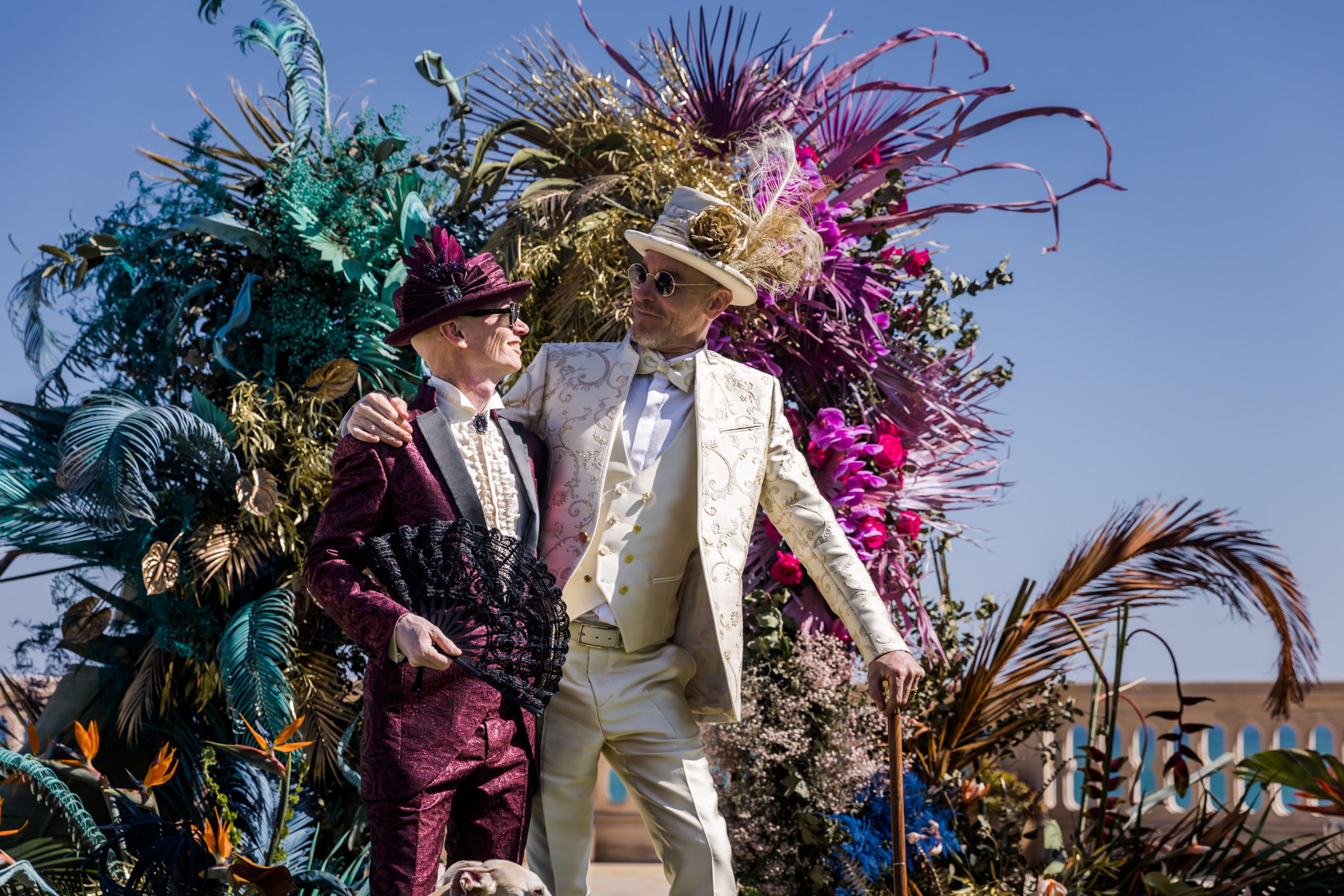 Adam Mason and Salvador Costa on their wedding day at Xalet del Nin, in front of a large floral installation against a blue sky