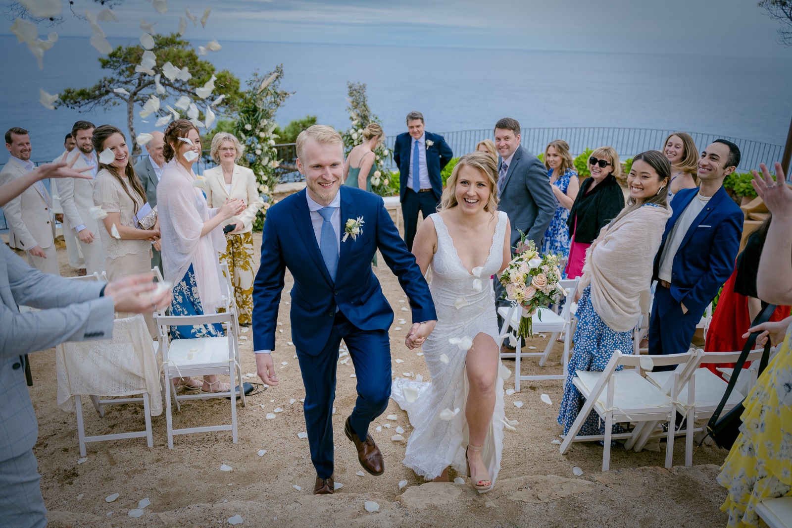 The bride and groom walk hand in hand up the aisle smiling as guests throw flower petals, with the ocean behind them.