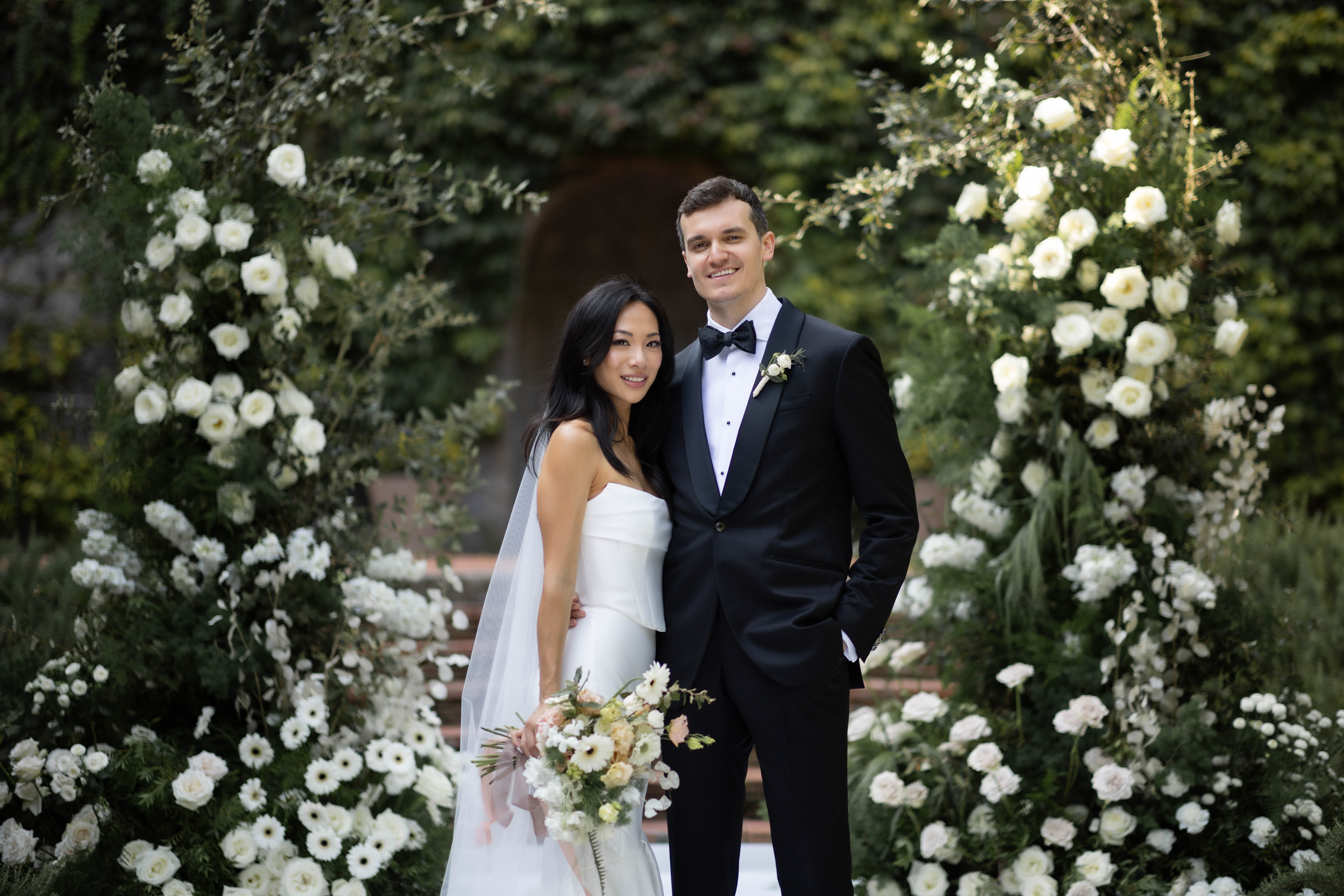 The bride and groom smile together in front of a large arch of white roses.