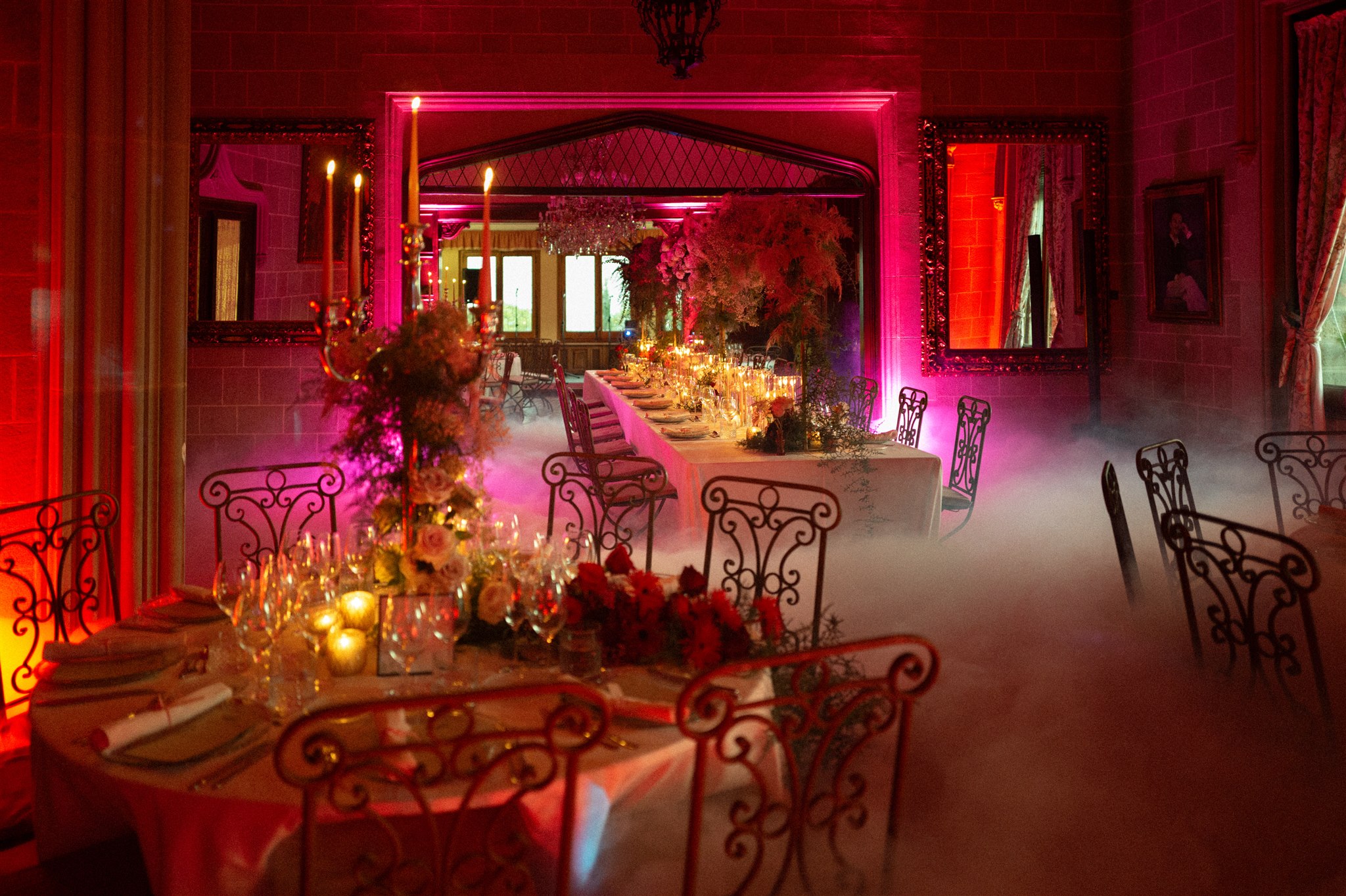 A long banquet table set for a wedding reception with a floral runner, tall candles, and pink uplighting in an ornate dining hall.