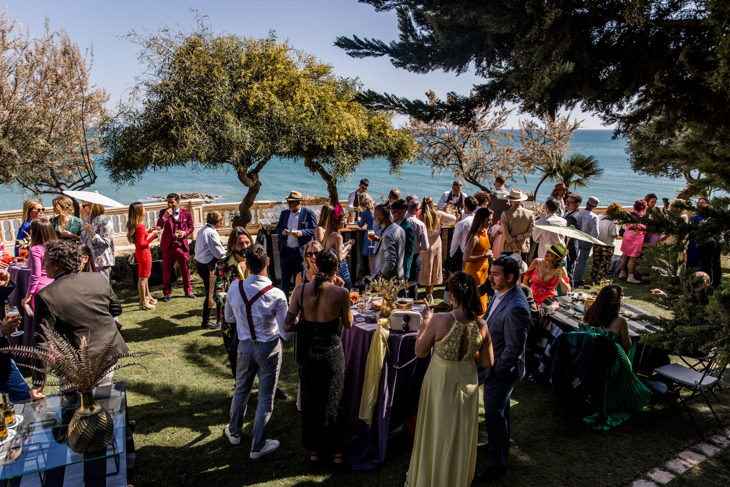 Guests at the aperitif at Xalet del Nin, standing with drinks on the lawn beneath the pines, the balustrade and the Mediterranean behind them