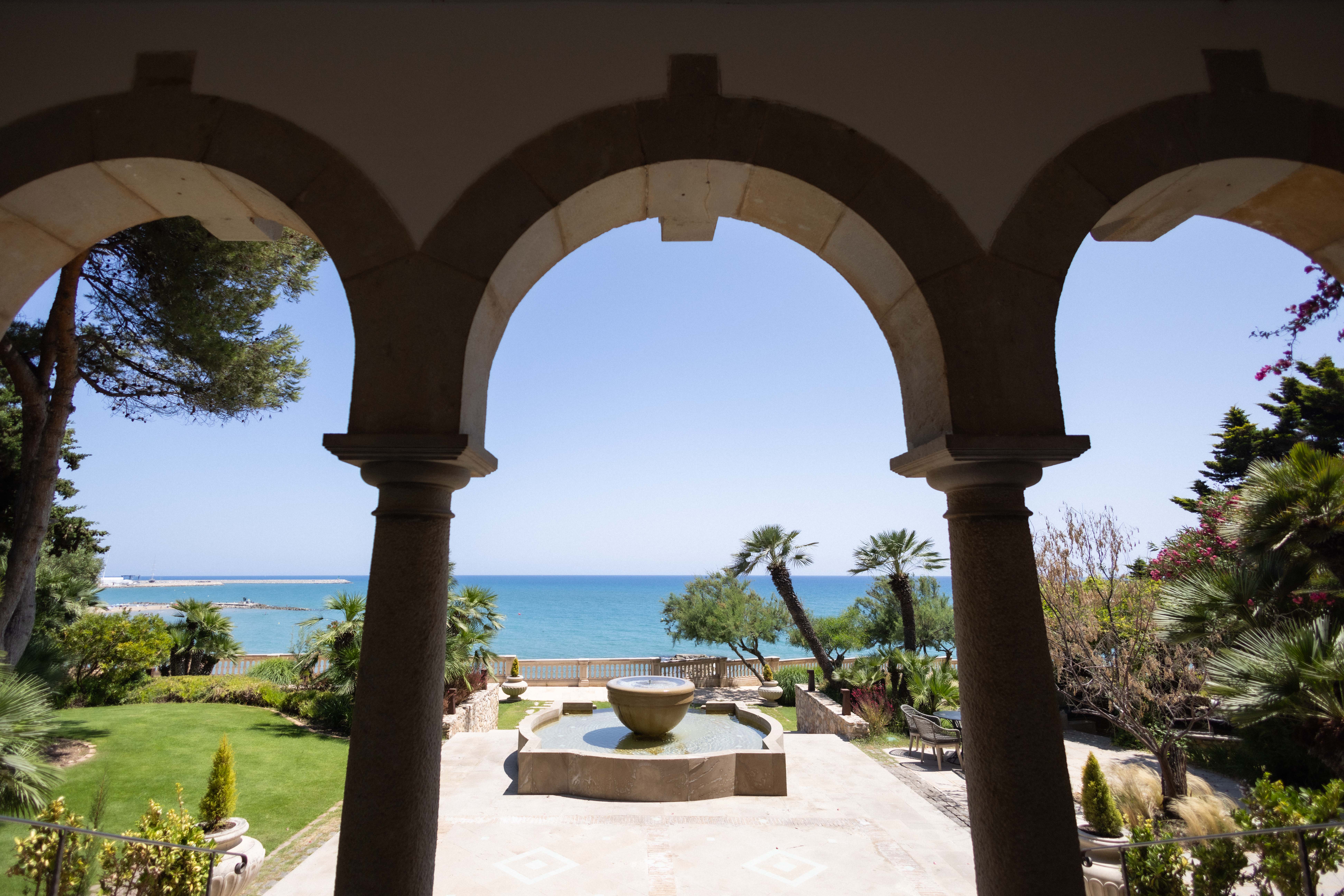 View through the stone arches at the front of Xalet del Nin, over the fountain and lawn to the Mediterranean