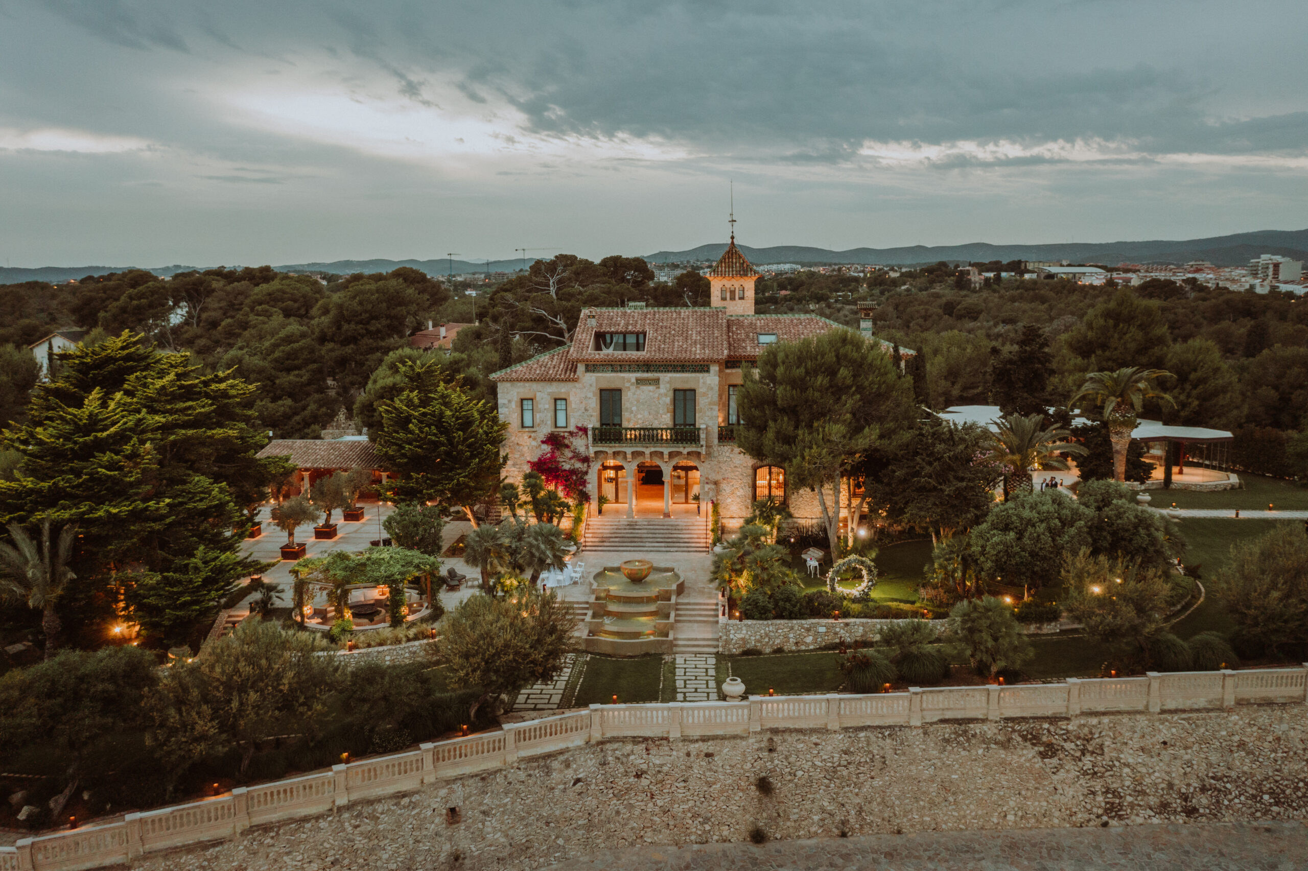 Xalet del Nin from above at dusk, the house lit and the gardens and fountain below, with the sea wall along the front